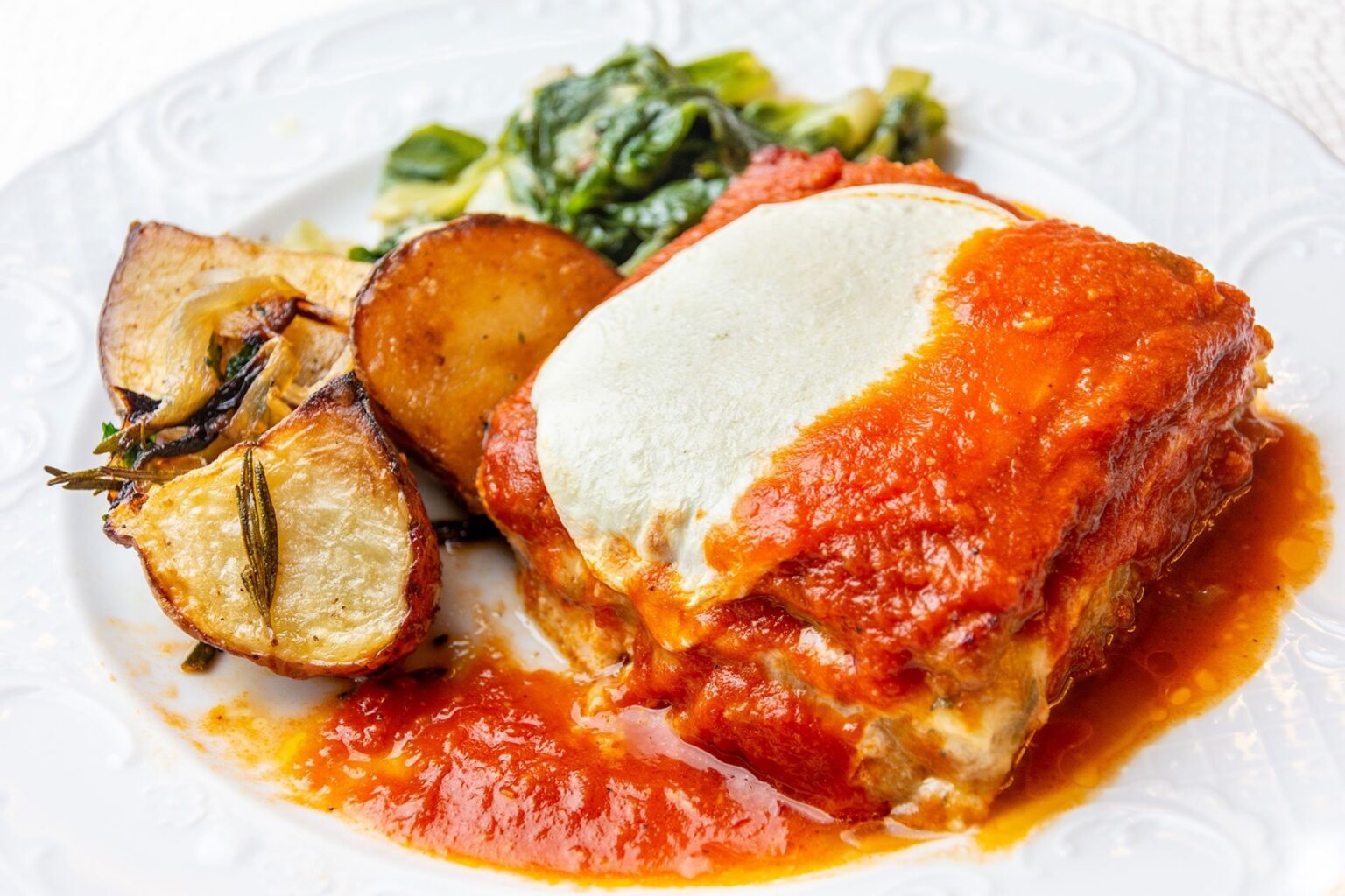 A layered baked dish with tomato sauce and melted mozzarella, roasted potatoes and greens