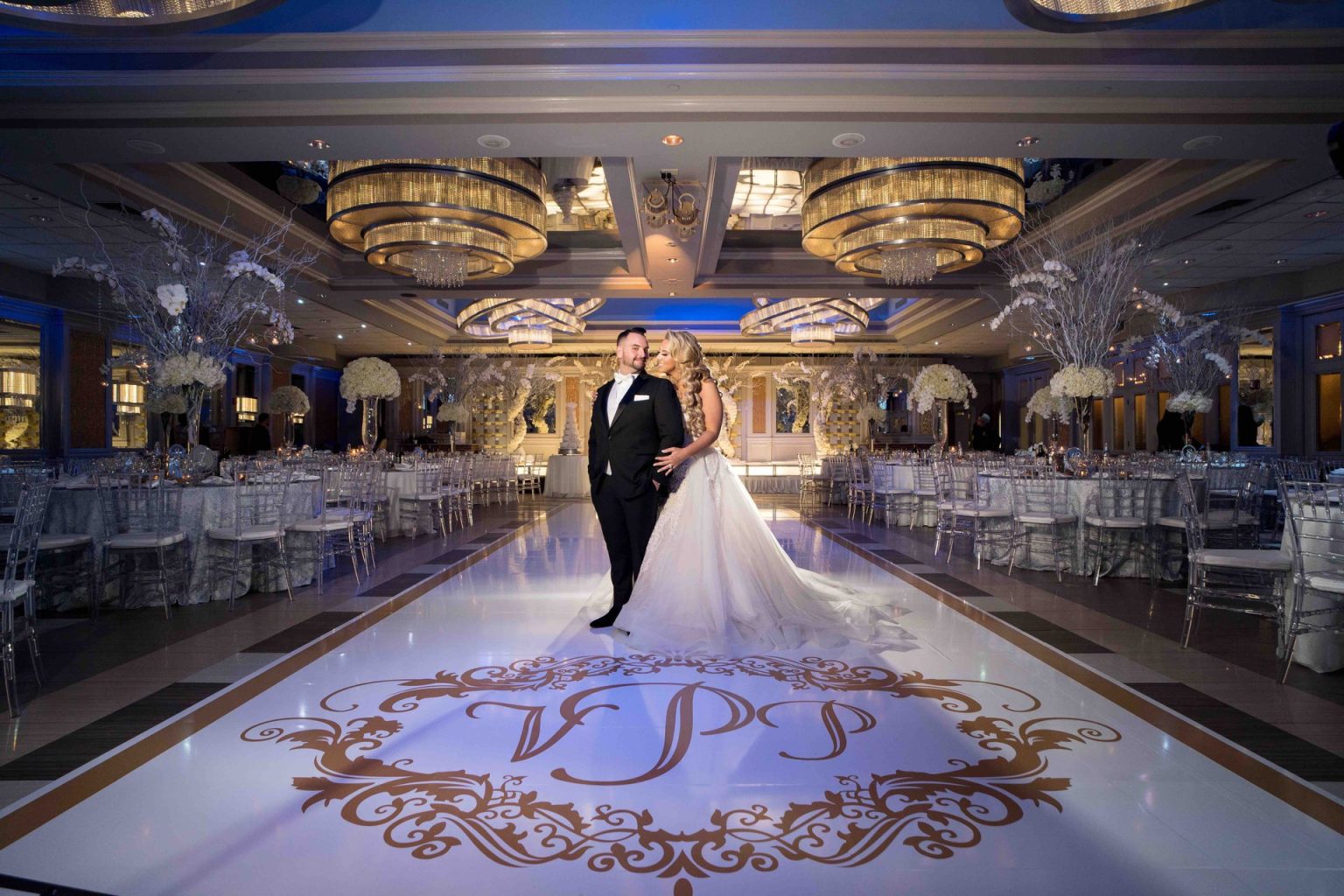A couple stands on their gold floor monogram in the Encore Ballroom, lit in blue