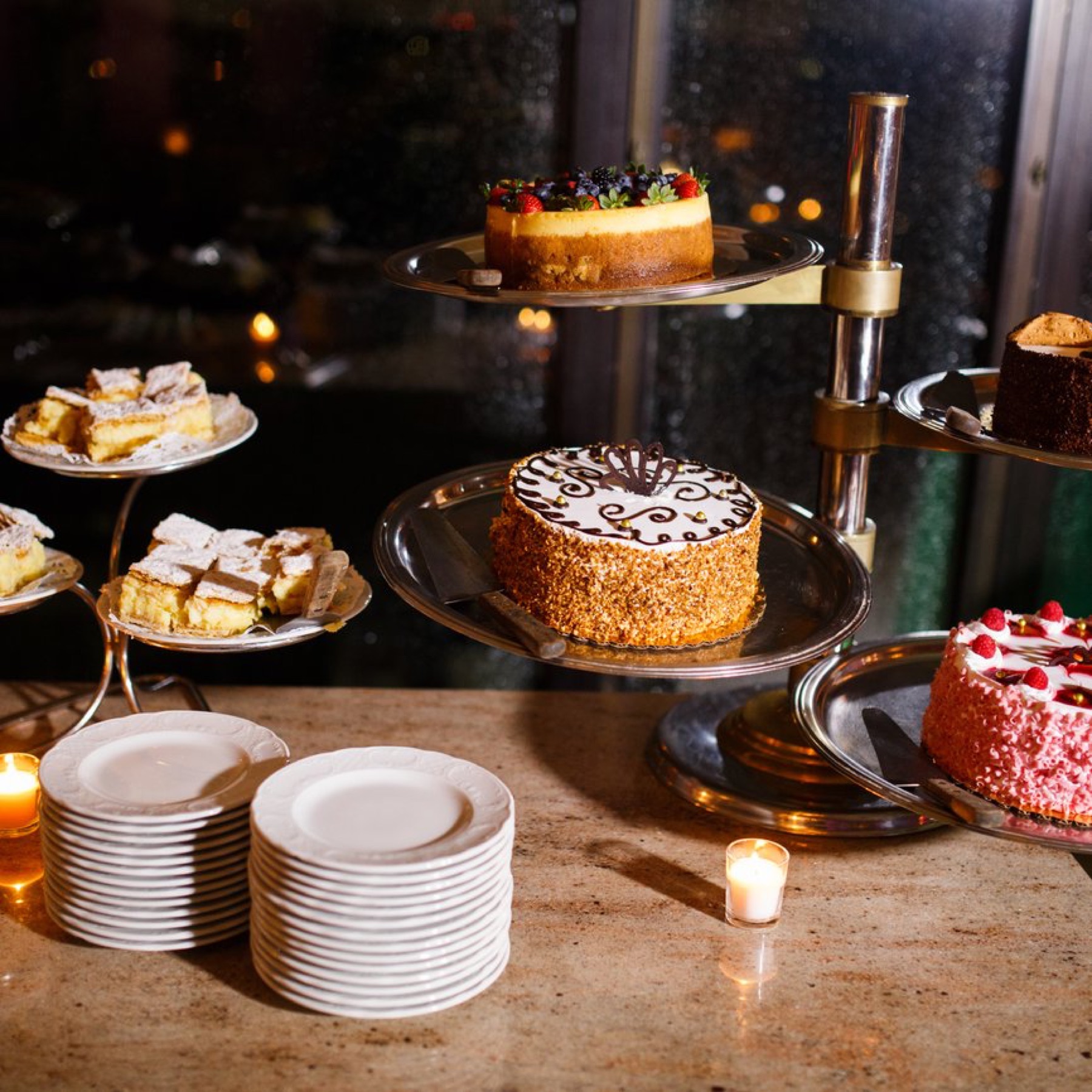Cakes and pastries on tiered stands by candlelight, with stacks of dessert plates