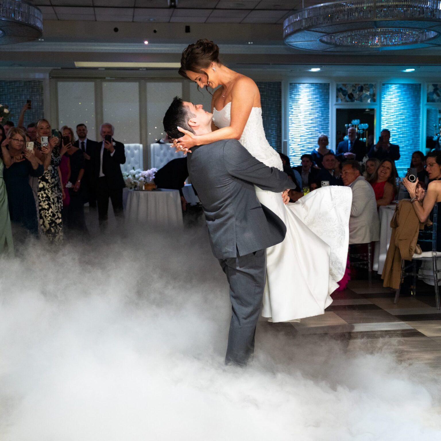 A groom lifts his bride during their first dance over low fog as guests watch