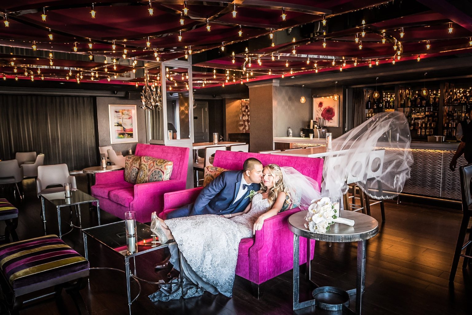 A bride and groom on a Rose Lounge sofa, her veil in the air, the bar behind